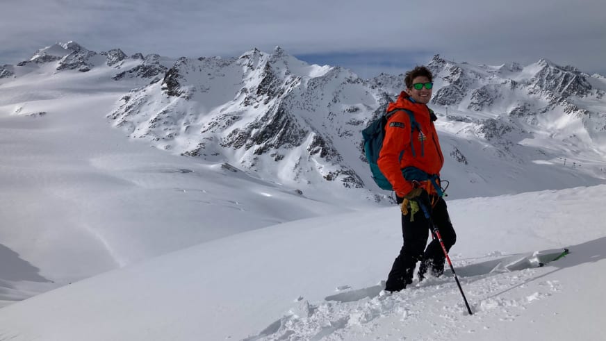 Am verschneiten Berggipfel steht Bergführer Korbinian Schmidtner in roter Jacke mit Skistock und blickt über das Hochgebirge.