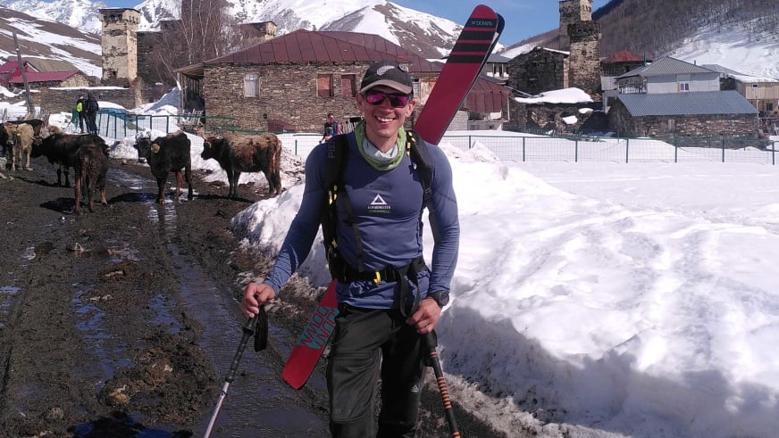 Bergführer Korbinian Schmidtner steht mit Skiausrüstung vor braunen Kühen in einem alpinen Dorf mit Steinhäusern.