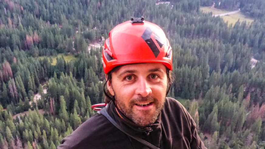 Bergführer Markus Hofmann macht ein Selfie mit rotem Helm am Gruenstein Klettersteig mit Waldblick im Hintergrund.