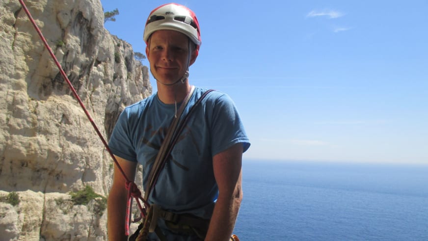 Bergführer Thomas Hochrein trägt rot-weißen Helm und blaues T-Shirt vor Kalksteinwand mit blauem Meer im Hintergrund.
