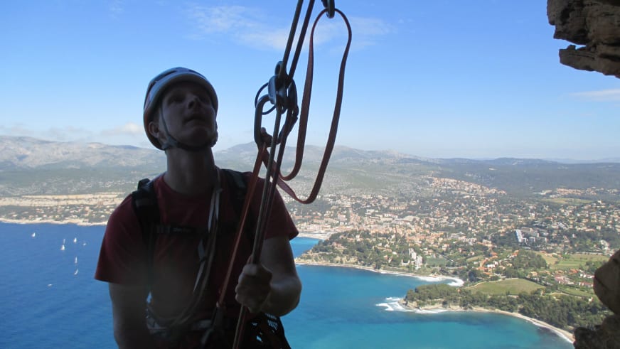 An einer Felswand hält Bergführer Thomas Hochrein Kletterseile vor mediteraner Küstenlandschaft mit türkisblauem Meer.