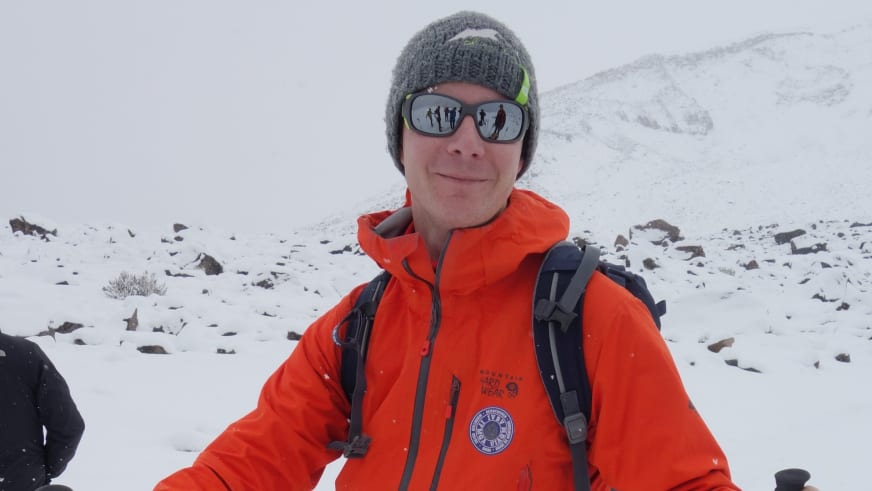 Bergführer Thomas Hochrein lächelt in die Kamera und trägt orange Winterjacke mit Kapuze vor schneebedeckter Berglandschaft im Vinschgau.