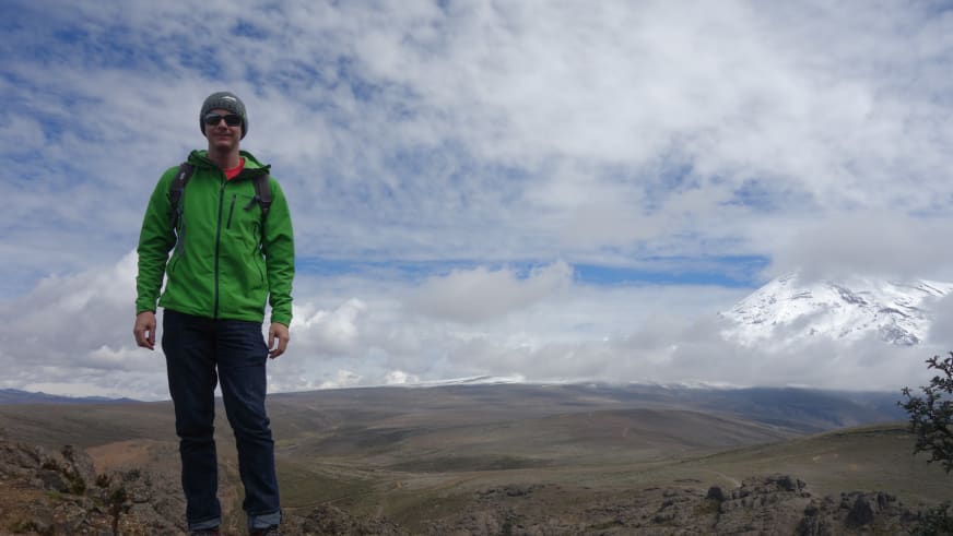 Bergführer Thomas Hochrein in grüner Softshell-Jacke und Wollmütze vor karger Hochlandschaft mit schneebedecktem Berggipfel.