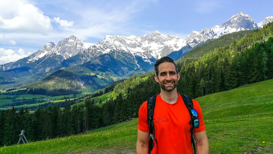 Bergwanderführer Alexander Plotzki lächelt vor schneebedeckten Felsgipfeln und trägt rotes T-Shirt mit schwarzem Rucksack.