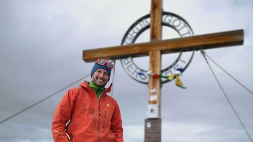 Am Gipfelkreuz mit bunten Gebetsfähnchen steht Bergführer Andreas Strasser in oranger Softshelljacke im dichten Nebel.