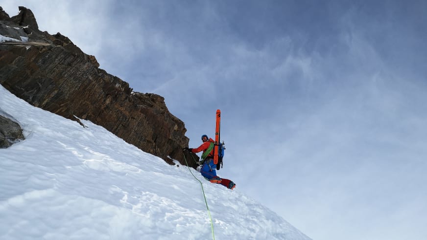 Einen steilen Schneehang steigt Bergführer Andreas Strasser in roter Jacke mit orangenen Skiern am Rücken und grünem Seil hinauf.