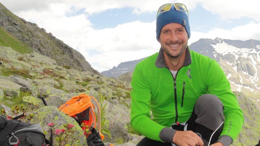 Zwischen Felsblöcken und rosa Bergblüten sitzt Bergführer Andreas Strasser lächelnd mit grüner Softshell-Jacke und blauer Mütze.