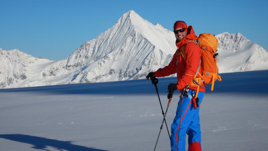 Bergführer Andreas Strasser steht in roter Winterjacke mit schwarzen Skistöcken und orangem Rucksack auf verschneiter Ebene.