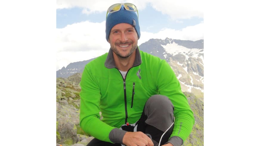 Bergführer Andreas Strasser sitzt entspannt auf steinigem Bergterrain mit blauer Strickmütze und grüner Softshell-Jacke.