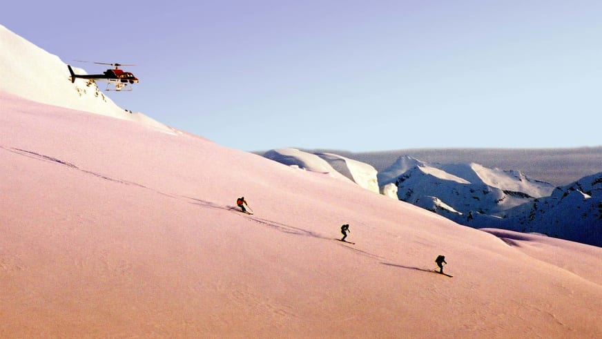 Bergführer Hubert Walther begleitet drei Skifahrer auf unverspurtem Schneehang, während rot-schwarzer Hubschrauber über ihnen schwebt.
