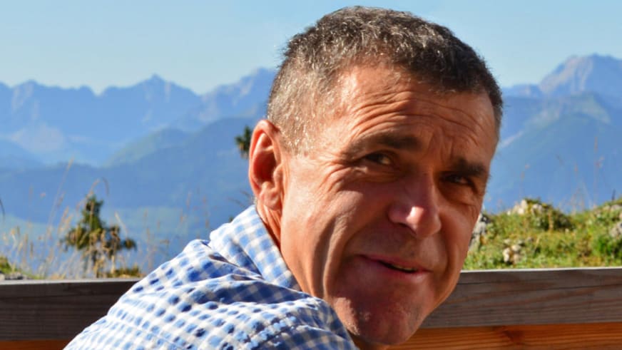 Portrait von Bergführer Hubert Walther in kariertem Hemd vor Bergpanorama auf einer Holzterrasse.