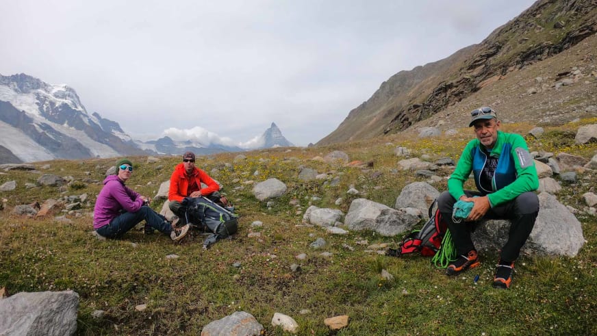 Bergführer Hubert Walther pausiert mit zwei Personen auf alpiner Wiese vor schneebedeckten Gipfeln.