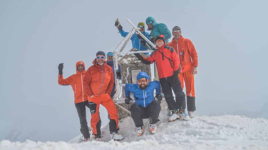 Acht Bergsteiger mit Bergführer Hubert Walther posieren am weißen Gipfelkreuz im verschneiten Hochgebirge.