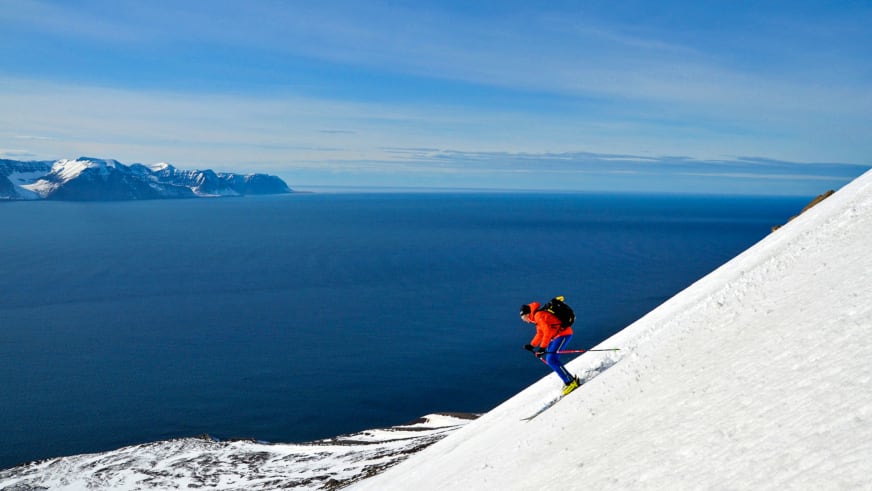 Bergführer Hubert Walther fährt in oranger Skijacke einen steilen verschneiten Hang mit Fjordblick hinab.