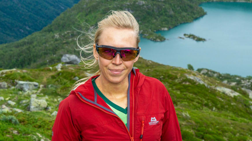 Bergwanderführerin Maria Müller mit roter Outdoorjacke und Sonnenbrille vor türkisblauem Wasser und bewaldeten Hügeln.