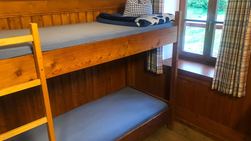 Schlafzimmer im Stöhrhaus mit Holz-Etagenbett, blauer Bettwäsche und Fenster mit kariertem Vorhang.
