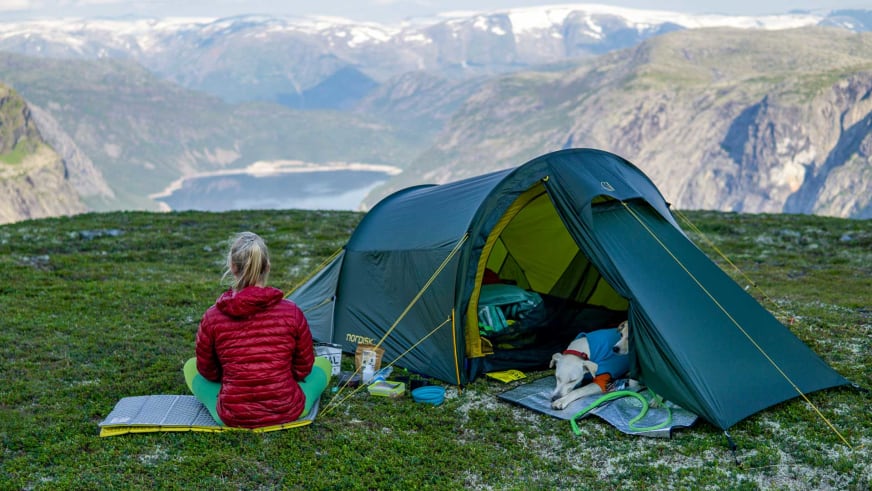 Bergwanderführerin Maria Müller in roter Daunenjacke sitzt auf Isomatte vor grünem Zelt mit Fjordlandschaft im Hintergrund.