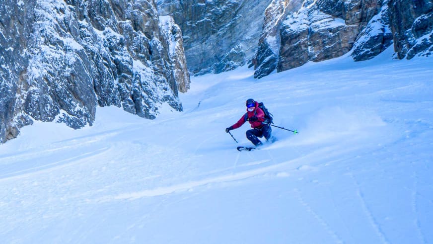 Bergwanderführerin Maria Müller fährt auf Skiern einen steilen Hang mit tiefem Pulverschnee ab, Schneewolken wirbeln um die Skier.