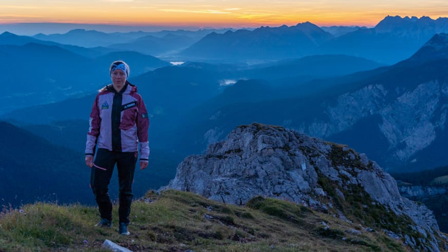 Bergwanderführerin Maria Müller steht auf Gipfelplateau mit Kalksteinblock vor weitläufigem Alpenpanorama bei goldener Lichtstimmung.