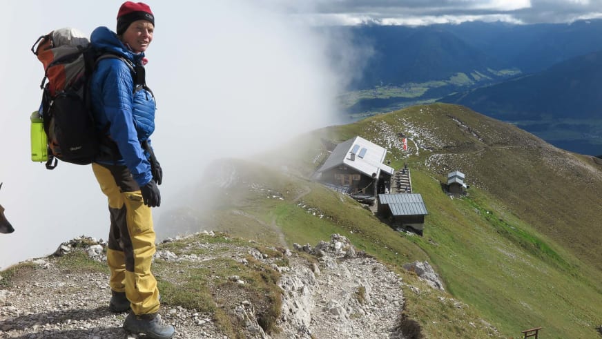 Mit blauer Daunenjacke und roter Mütze steht Bergwanderführerin Andrea Beuchlen auf einem felsigen Grat vor einer Berghütte.