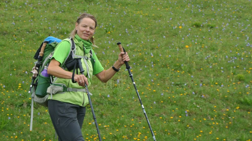 In blühender Almwiese mit gelben und violetten Wildblumen steht Bergwanderführerin Andrea Beuchlen mit grünem Rucksack und Wanderstöcken.