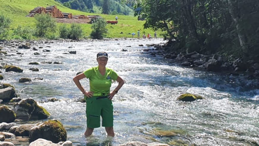 Bergwanderführerin Andrea Beuchlen steht mit grünem T-Shirt und Wanderhose in einem steinigen Bergbach bei hellem Tageslicht.