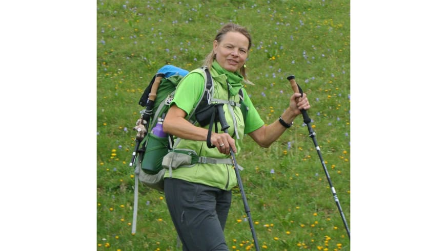Bergwanderführerin Andrea Beuchlen lächelt in die Kamera und trägt grünes Funktionsshirt mit Wanderrucksack auf einer Blumenwiese.