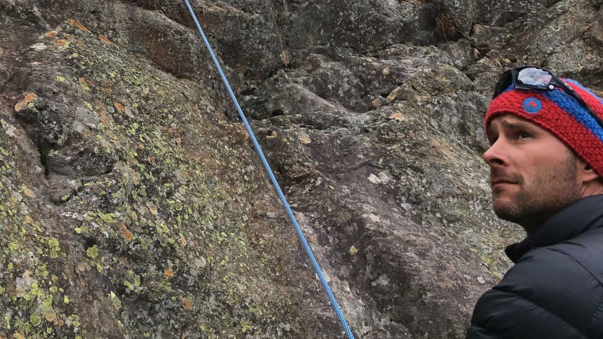 An einer Felswand mit gelblich-grünen Flechten steht Bergführer Uwe Berg in rot-blau-gestreifter Mütze und grauer Softshell-Jacke.