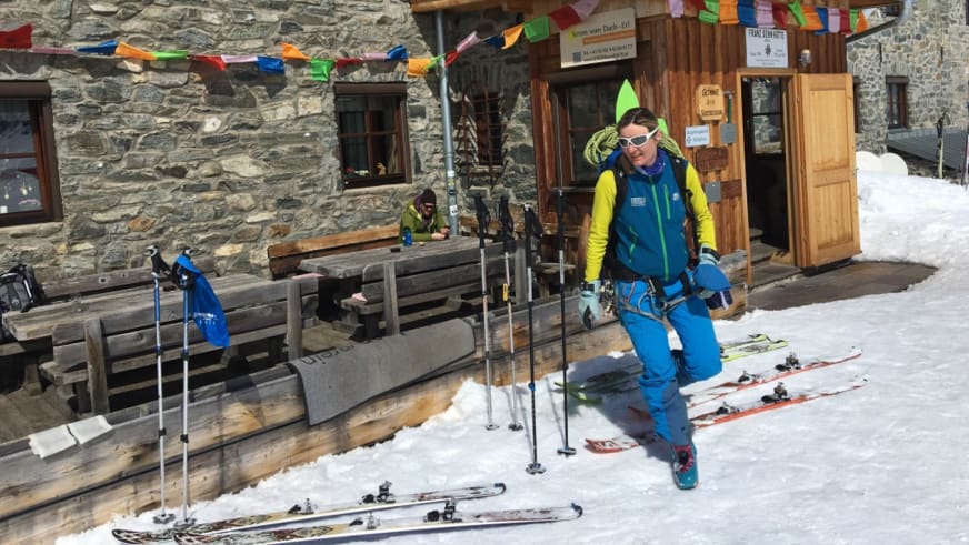 An einer alpinen Berghütte mit Natursteinwänden schnallt Bergwanderführerin Karin Toni Bigel ihre Tourenskier ab.