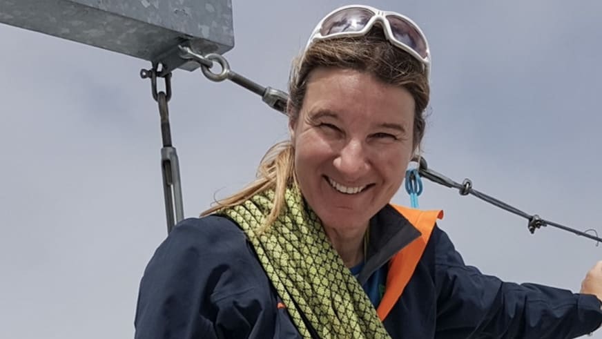 Bergwanderführerin Karin Toni Bigel lächelt freundlich vor einer Seilbahnkonstruktion und trägt ein gelbes Kletterseil um den Hals.