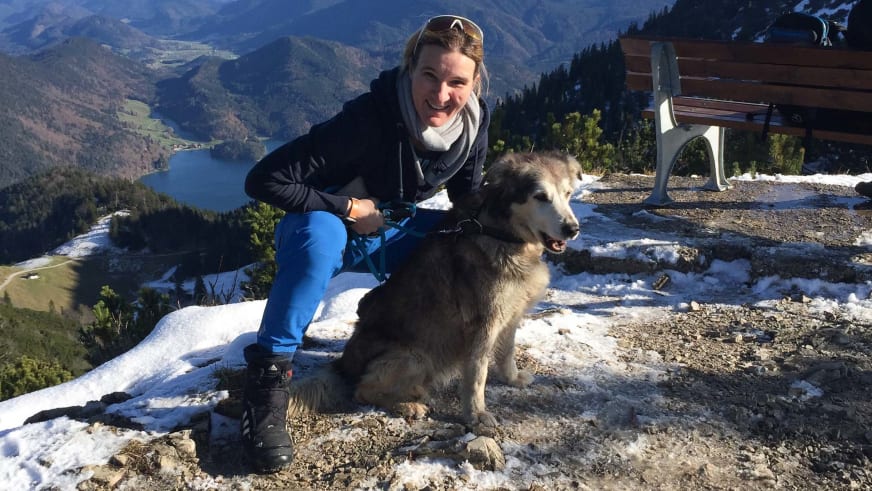 Auf einem schneebedeckten Aussichtspunkt sitzt Bergwanderführerin Karin Toni Bigel neben einem grau-weißen Hund.