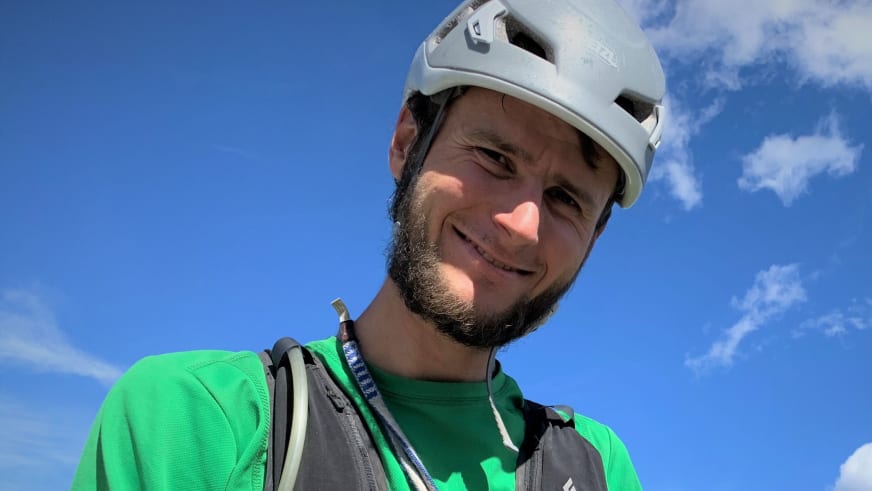 Portrait von Bergführer Jonas Wolkenstörfer mit weißem Kletterhelm und grünem Shirt vor blauem Himmel.