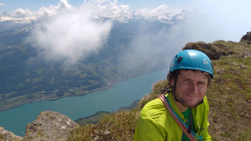 Auf exponiertem Berghang steht Bergführer Jonas Wolkenstörfer mit blauem Helm und türkisfarbenem See im Hintergrund.