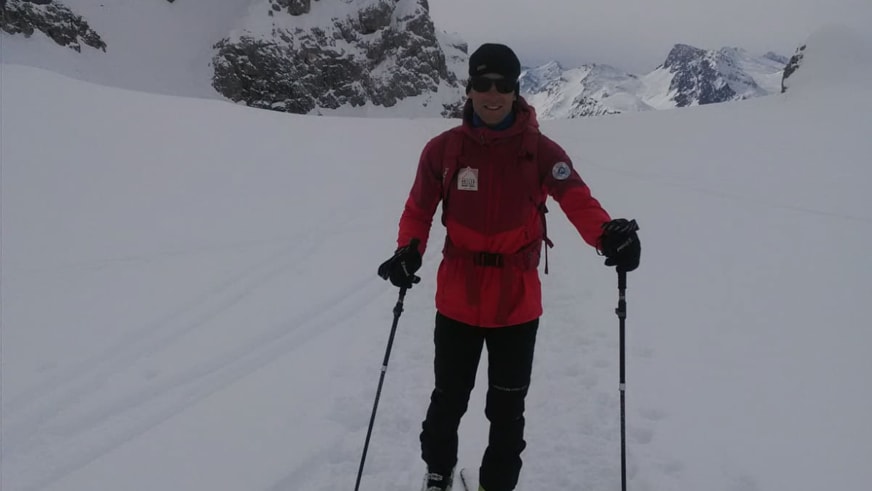 Bergführer Rolando Varesco steht lächelnd mit Skistöcken und roter Winterjacke vor verschneiten Berggipfeln.