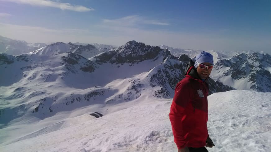 Lächelnd steht Bergführer Rolando Varesco in roter Winterjacke auf schneebedecktem Gipfel vor Hochgebirgspanorama.