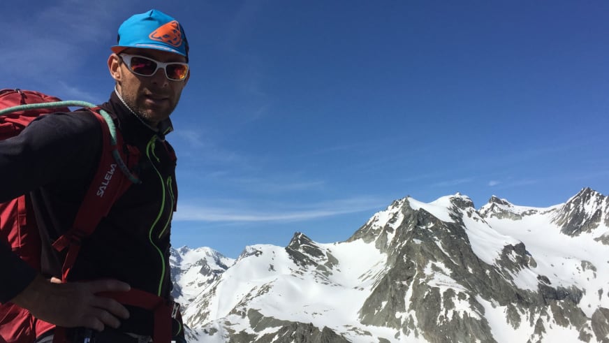 Bergführer Rolando Varesco trägt blaue Mütze mit orangem Logo und schwarze Softshelljacke vor verschneiter Bergkette.