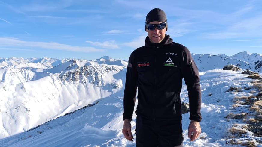 Auf einem schneebedeckten Gipfel steht Bergführer Rolando Varesco mit schwarzer Sonnenbrille vor verschneiten Berggipfeln.