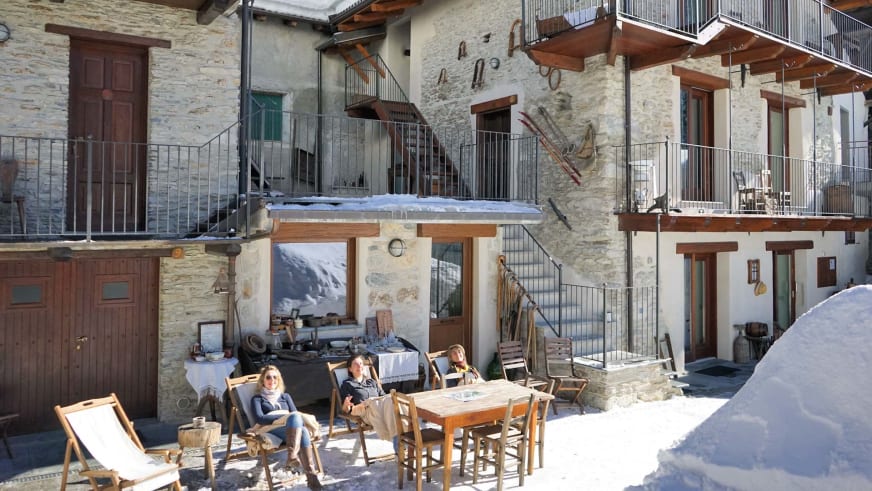 Sonnige Winterterrasse der Pensione Ceaglio Valle Maira mit Holzmöbeln und traditioneller Steinarchitektur im Innenhof.