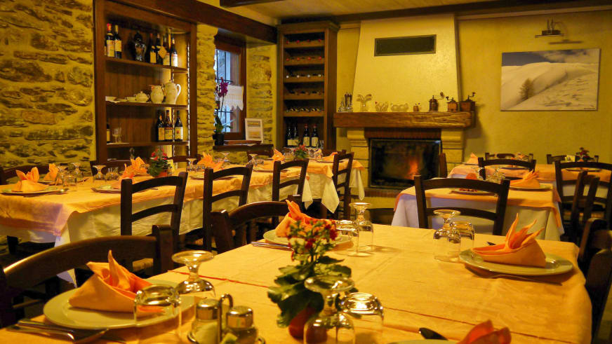 Restaurant der Pensione Ceaglio zeigt eingedeckte Holztische, brennenden Kamin und Natursteinmauerwerk im Valle Maira.