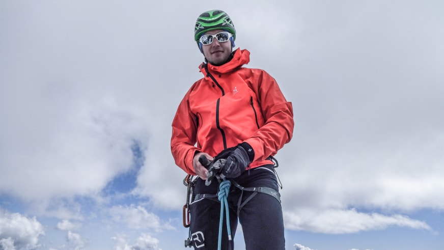 Mit roter Softshell-Jacke und Kletterausrüstung posiert Bergführer Matthias Hahn vor bewölktem Himmel in Froschperspektive.