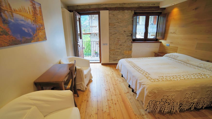 Zimmer Preit im Hotel Ceaglio mit Natursteinwand, Holzbalken an der Decke und zwei weißen Sesseln am Fenster.