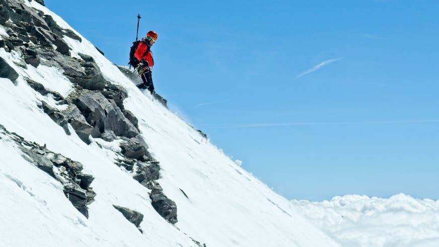 Bergführer Matthias Hahn in roter Winterjacke und orangenem Helm steigt einen steilen schneeverschneiten Hang mit Felseinlagerungen auf.