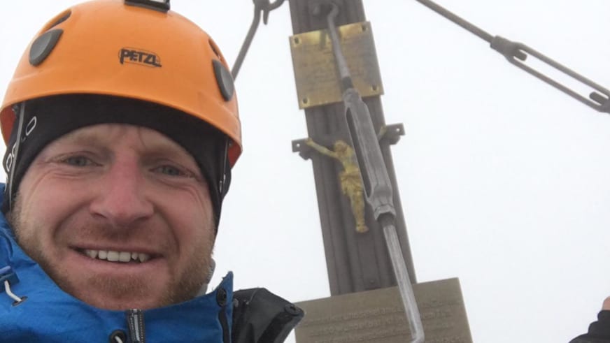 Bergführer Michael Schmidt lächelt am Gipfel mit orangem Petzl-Kletterhelm und blauer Funktionsjacke vor Gipfelkreuz.