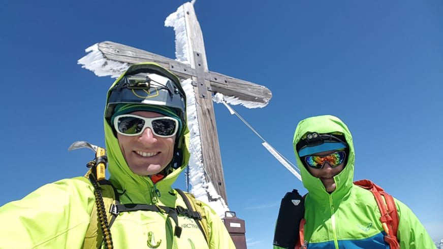 Bergführer Martin Riener steht mit zweiter Person vor verreiftem Holzkreuz auf Berggipfel bei klarem Winterwetter.