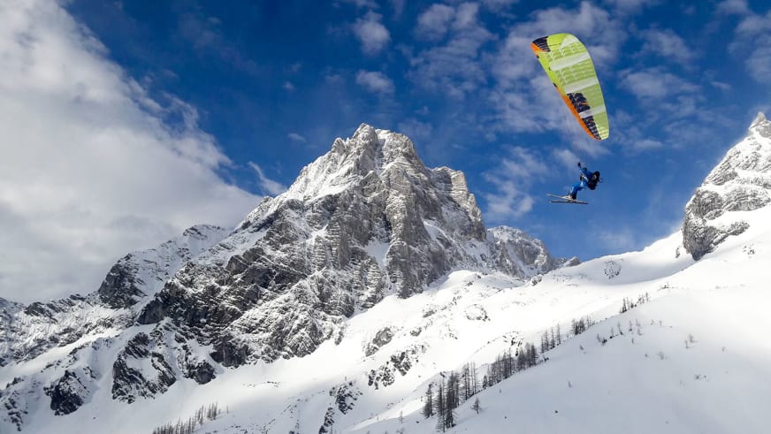 Bergführer Martin Riener schwebt mit grün-orange Gleitschirm und Skiern über verschneiter Berglandschaft bei klarem Himmel.