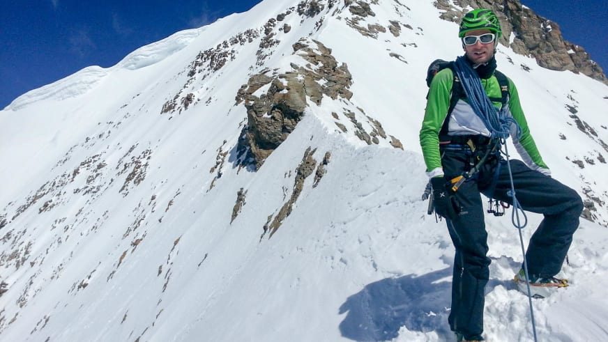 In grüner Softshelljacke und Steigeisen pausiert Bergführer Martin Riener vor verschneiter Felswand im Hochgebirge.