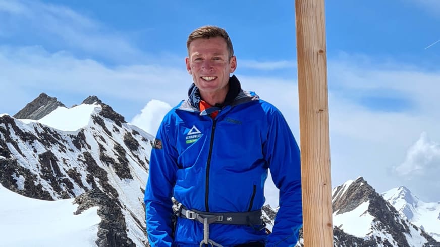 Bergführer Martin Riener in blauer Funktionsjacke mit Alpinewelt-Logo lächelt vor hölzernem Gipfelkreuz in schneebedeckter Hochgebirgslandschaft.