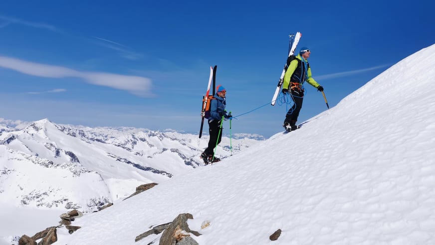 Zwei Skitourengeher mit Bergführer Martin Riener steigen durch verschneites Hochgebirge auf, Tourenskier auf dem Rücken tragend.