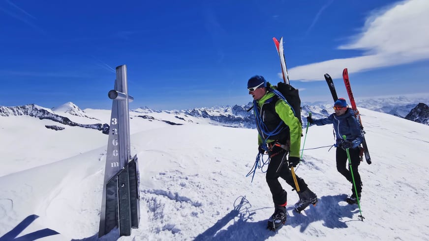 Bergführer Martin Riener in grüner Skijacke steht mit Partner am Gipfelkreuz eines schneereichen Berggipfels nach Skitour.
