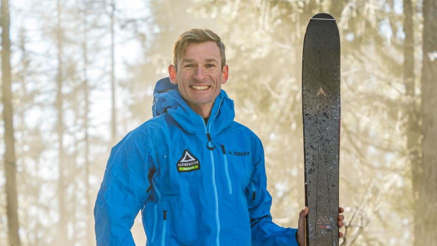 Bergführer Martin Riener lächelt in blauer Alpine Welten Jacke und hält grauen Tourenski vor kahlen Bäumen.